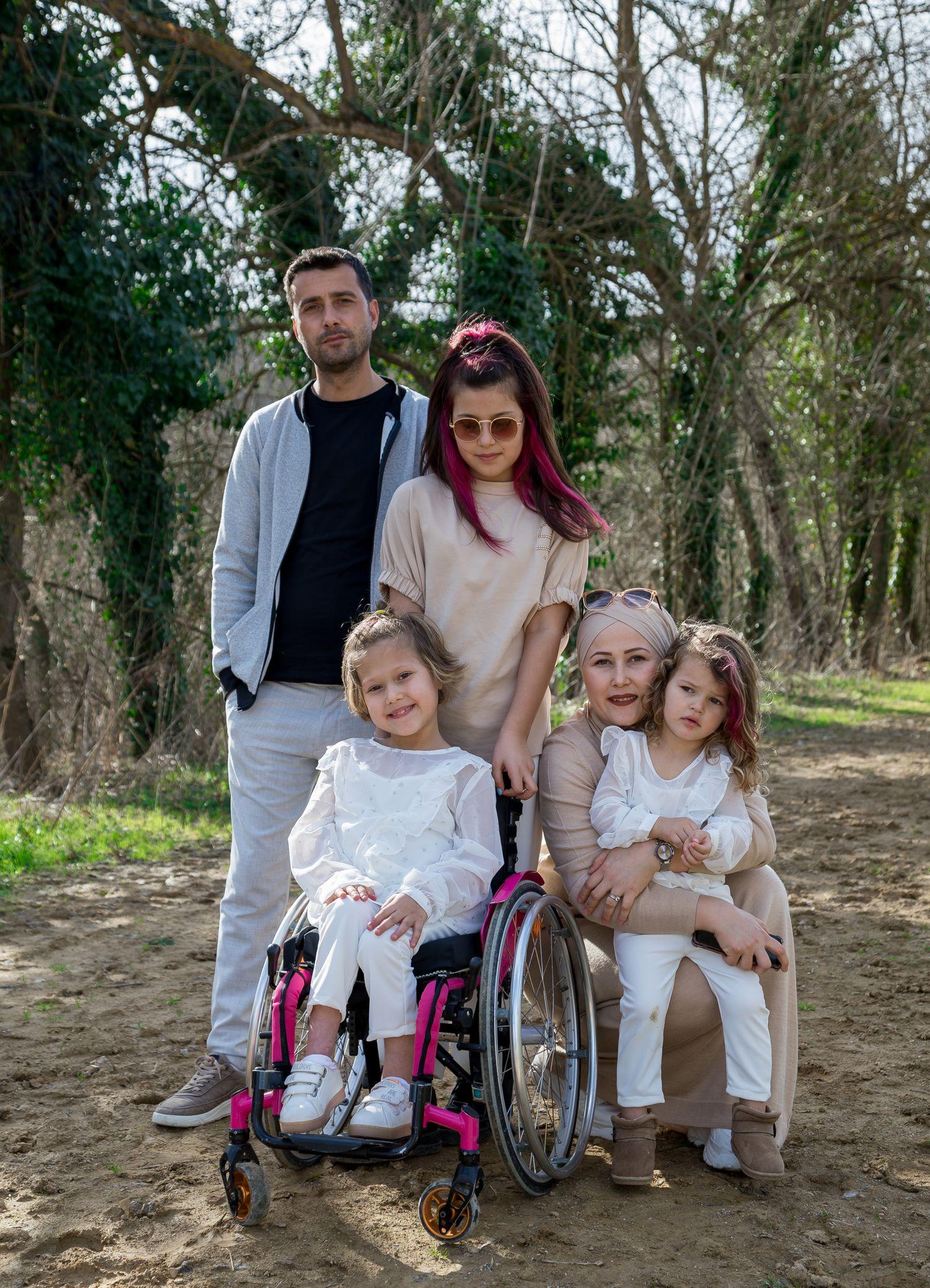 A family outdoors with their young daughter, who uses a wheelchair, gathered close together and smiling