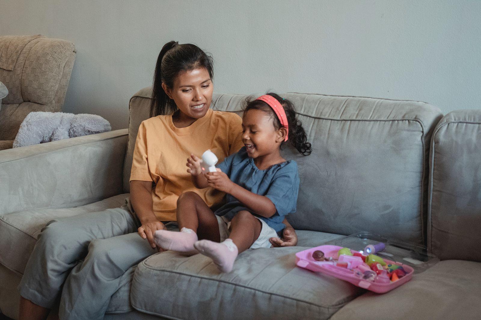 A mother smiling with her young daughter on the couch at home during a playful checkup with a toy medical kit